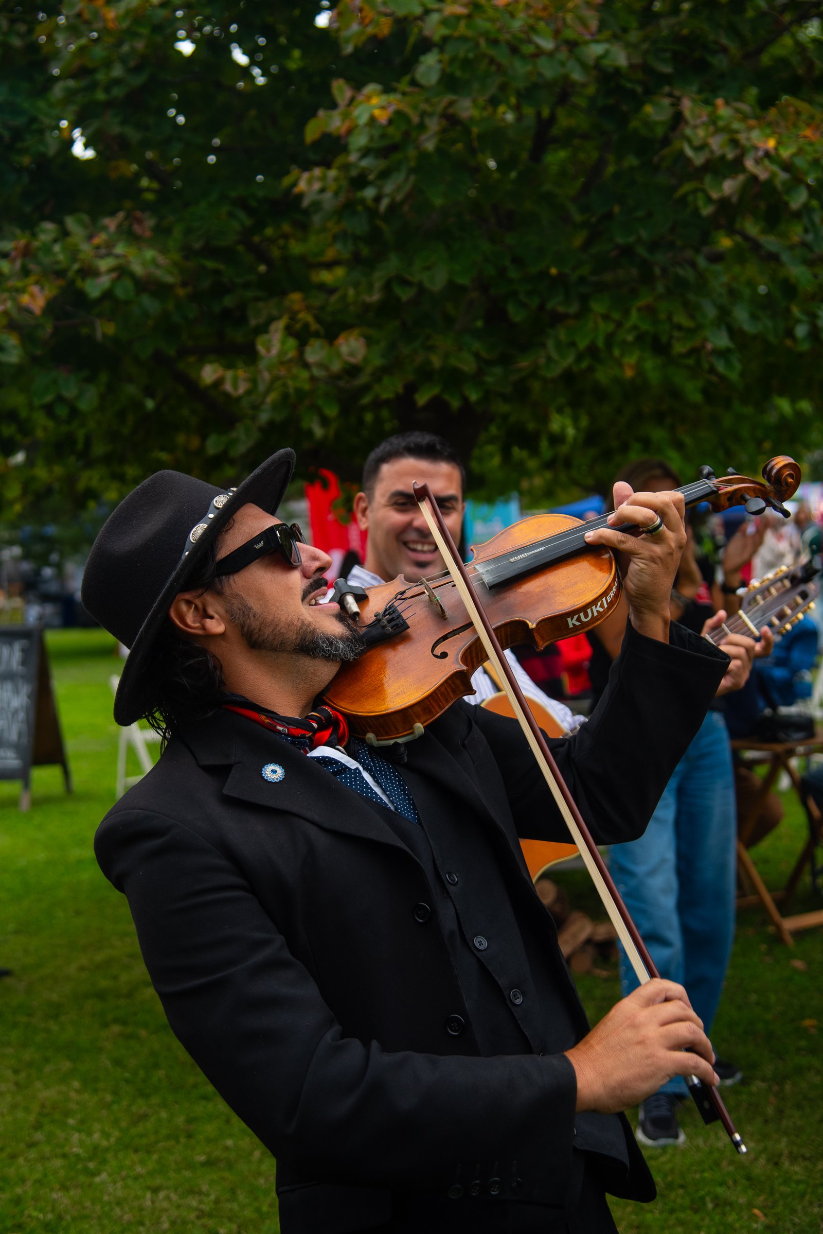 Violinista tocando en vivo con sombrero y anteojos de sol bajo los árboles